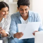 A smiling couple sitting together, reviewing documents and using a smartphone