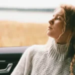 A woman is seated in the passenger side of a car, looking out the window at a serene field, reflecting on her thoughts or mental health in a moment of tranquility.