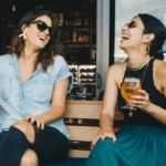 Two women sitting outdoors, laughing with a glass of beer in their hands