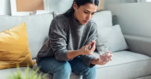 A woman sitting on a couch, holding a pill in one hand and a glass of water in the other, preparing to take prescription medication