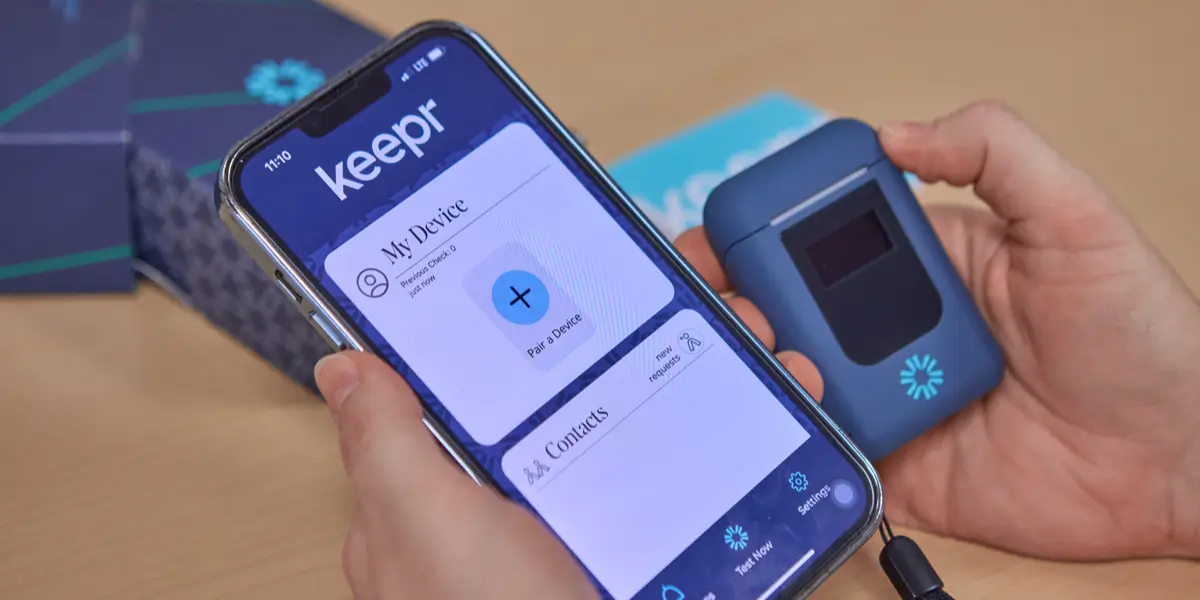 Person holding a smartphone displaying the Keepr app interface next to a compact Keepr personal breathalyzer device
