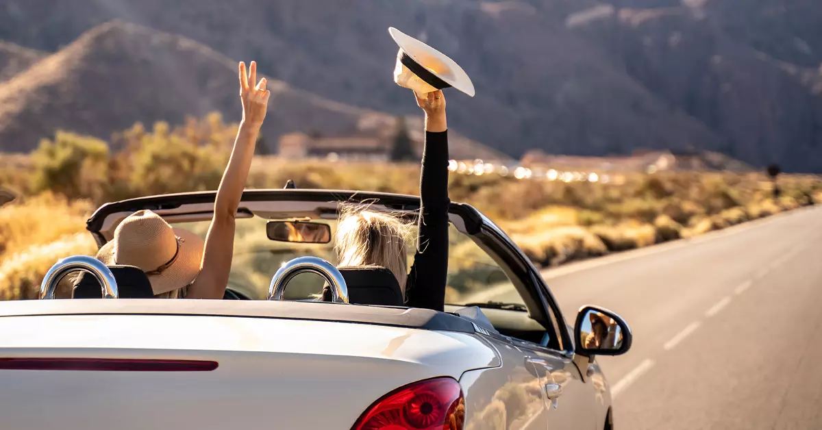 Two people enjoying a road trip in a convertible car, raising their hands in the air while driving through a scenic desert landscape.