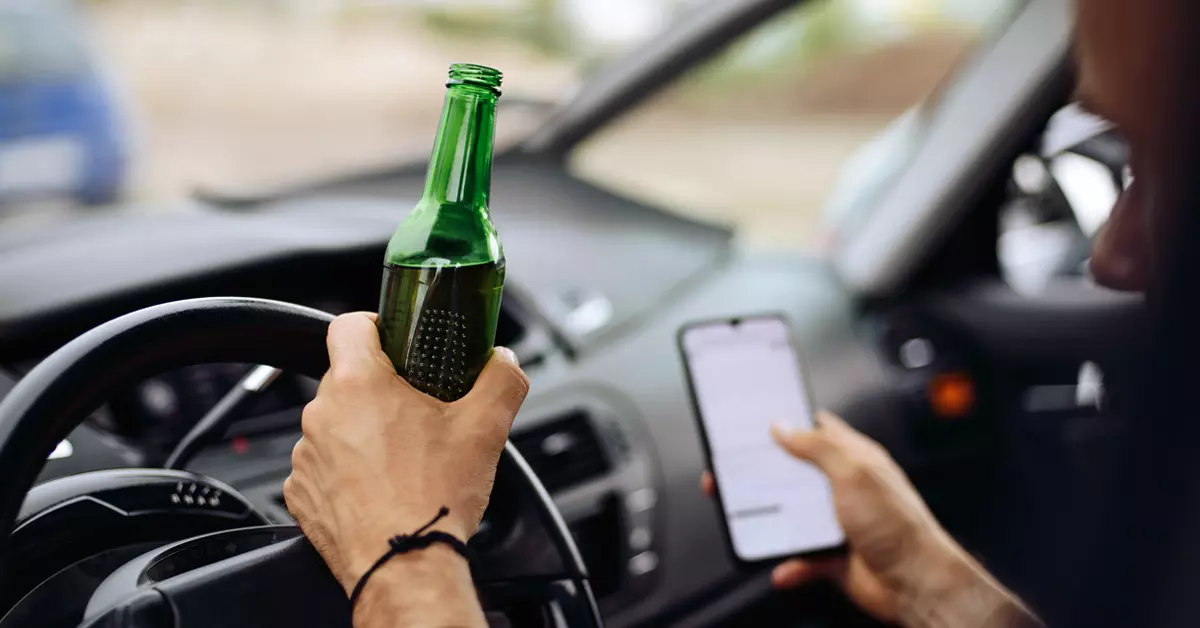 A closeup of a person holding a beer and a phone while driving a car.