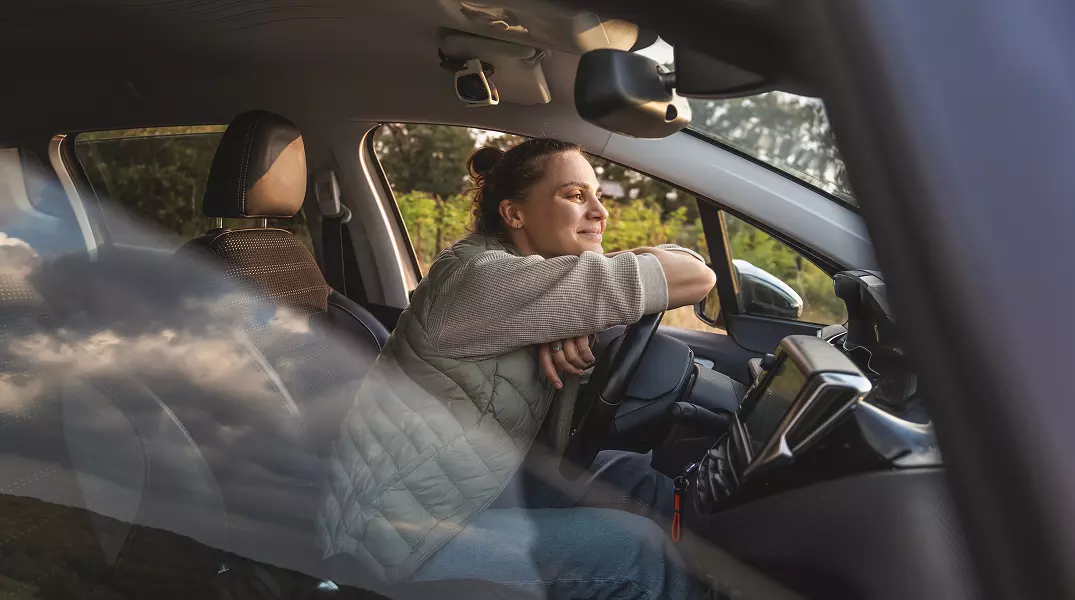 Woman resting on the steering wheel of her parked car looking forward and smiling.