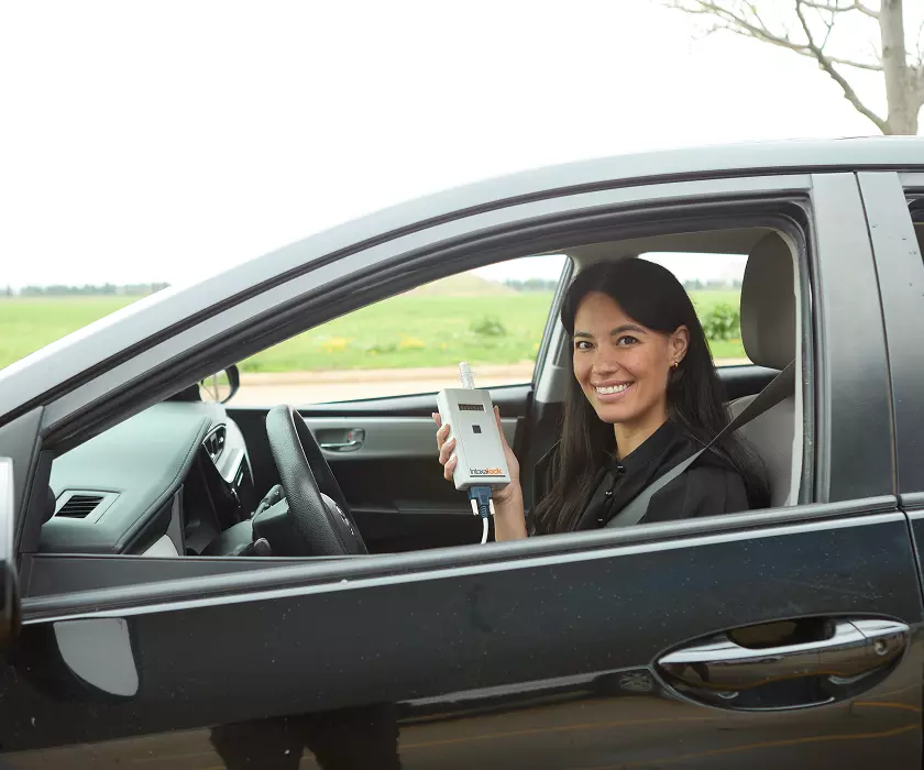 Woman smiling and sitting in her car while holding up Intoxalock IID device.